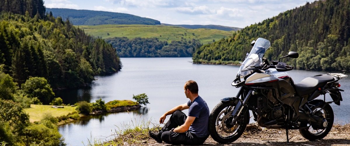 A man wearing black leather pants sitting on the ground next to a black adventure motorcycle on the edge of a cliff.