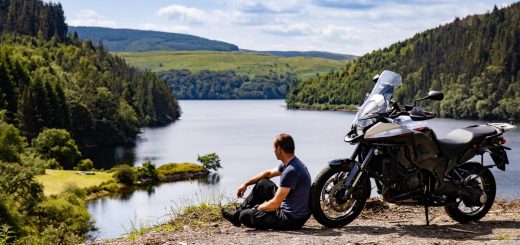 A man wearing black leather pants sitting on the ground next to a black adventure motorcycle on the edge of a cliff.