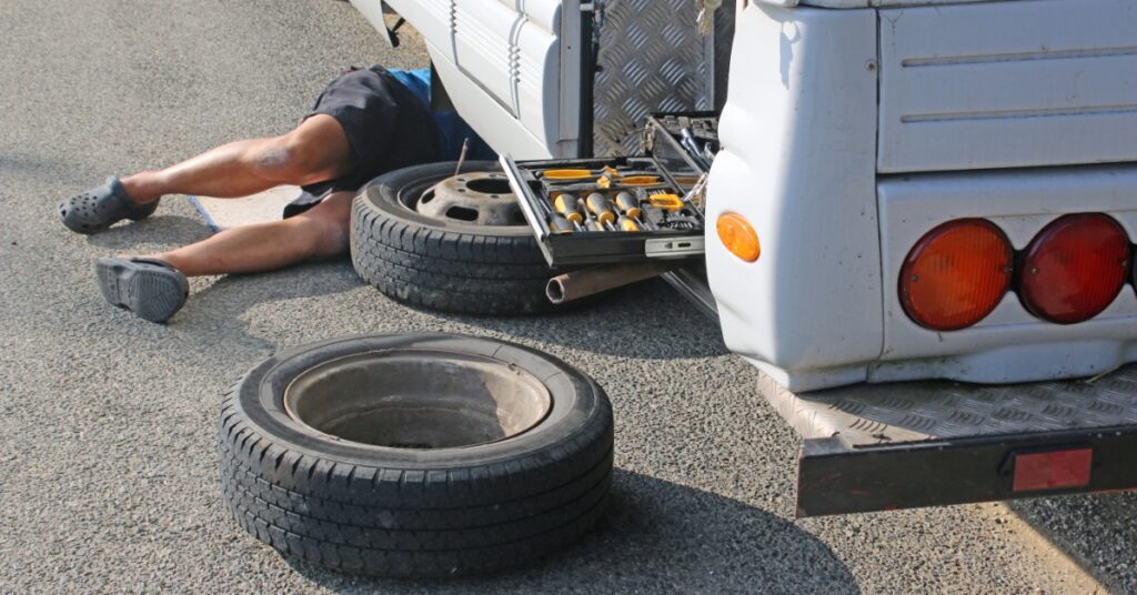A man is lying under an RV. There is a tire on the road and a tire missing from the RV’s wheel well.