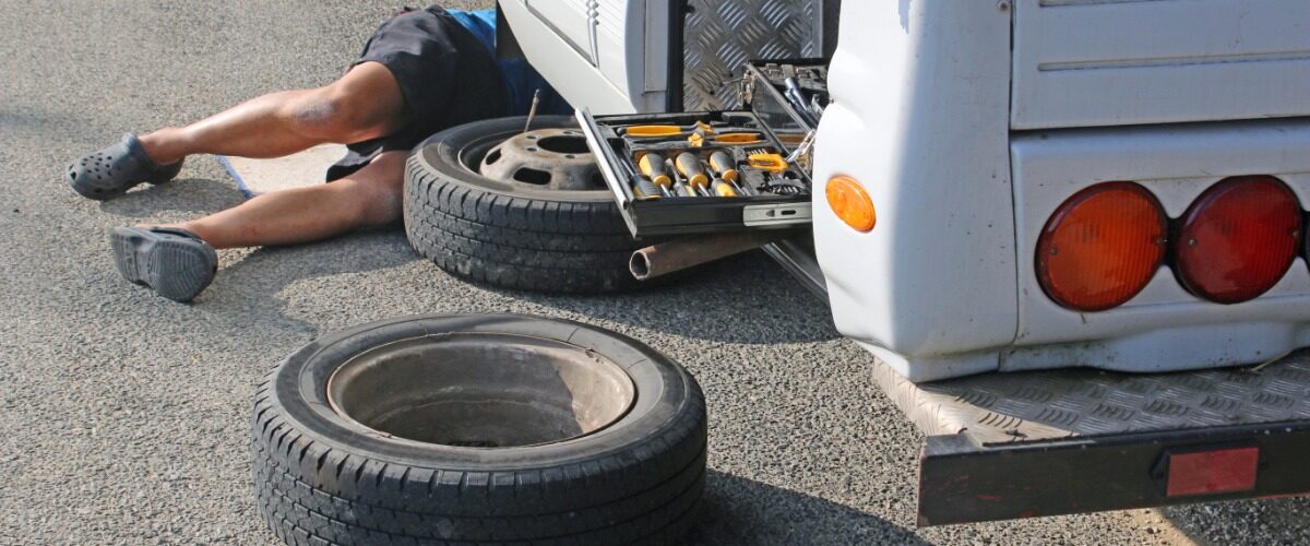 A man is lying under an RV. There is a tire on the road and a tire missing from the RV’s wheel well.
