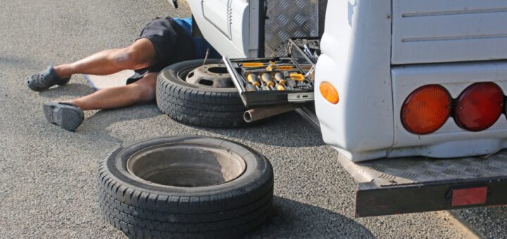 A man is lying under an RV. There is a tire on the road and a tire missing from the RV’s wheel well.