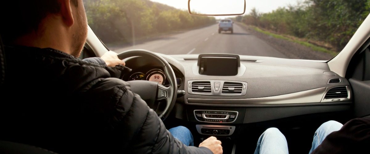 A man is sitting in the driver’s seat of a car. He’s driving down the road, and there is another car ahead of him.