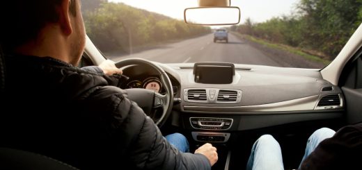 A man is sitting in the driver’s seat of a car. He’s driving down the road, and there is another car ahead of him.