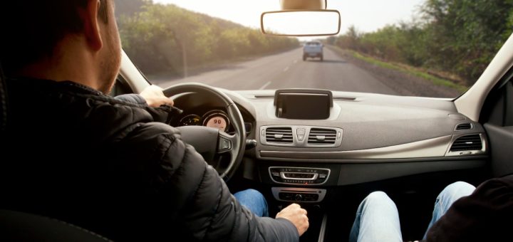 A man is sitting in the driver’s seat of a car. He’s driving down the road, and there is another car ahead of him.