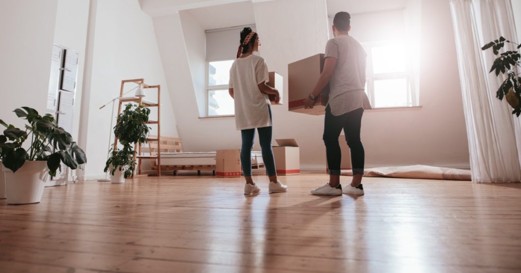 A man and a woman are standing in the middle of a living room. They are both holding boxes in their hands.