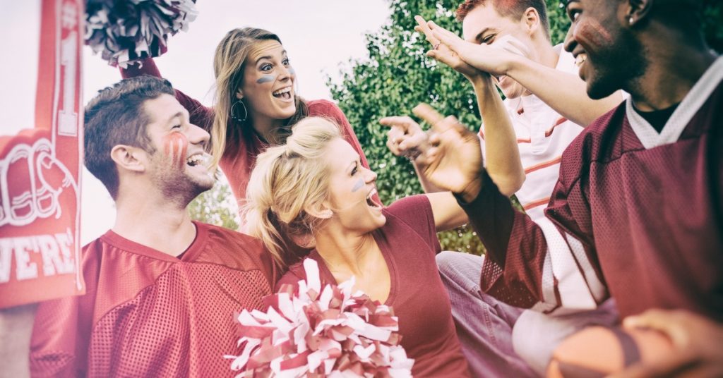 Group of college football fans in team colors cheering and high-fiving together at an outdoor tailgate.