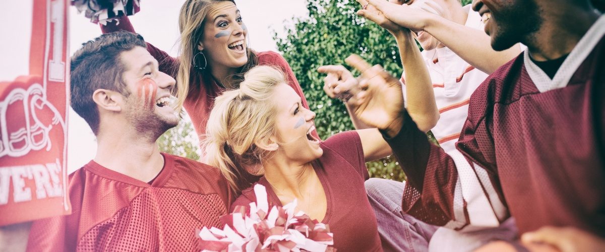 Group of college football fans in team colors cheering and high-fiving together at an outdoor tailgate.