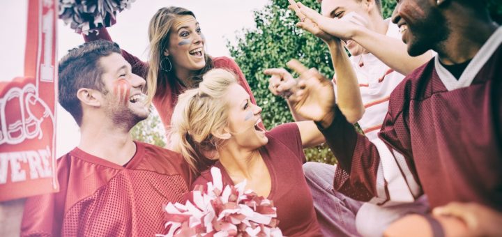 Group of college football fans in team colors cheering and high-fiving together at an outdoor tailgate.